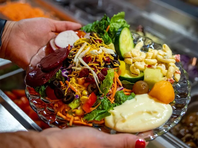 Hands holding a glass plate piled with colorful salad bar toppings, pasta salad, and creamy dressing