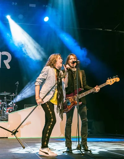 Two male rock musicians singing on a blue-lit stage, one playing a bass guitar