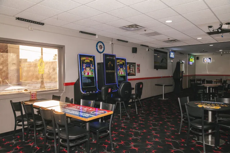 Dimly lit bar room with arcade machines, black chairs, patterned carpet, and wall-mounted TVs