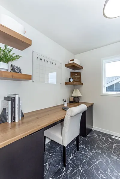 Home office with wood desk, white upholstered chair, floating shelves, and dark marble-look tile floor