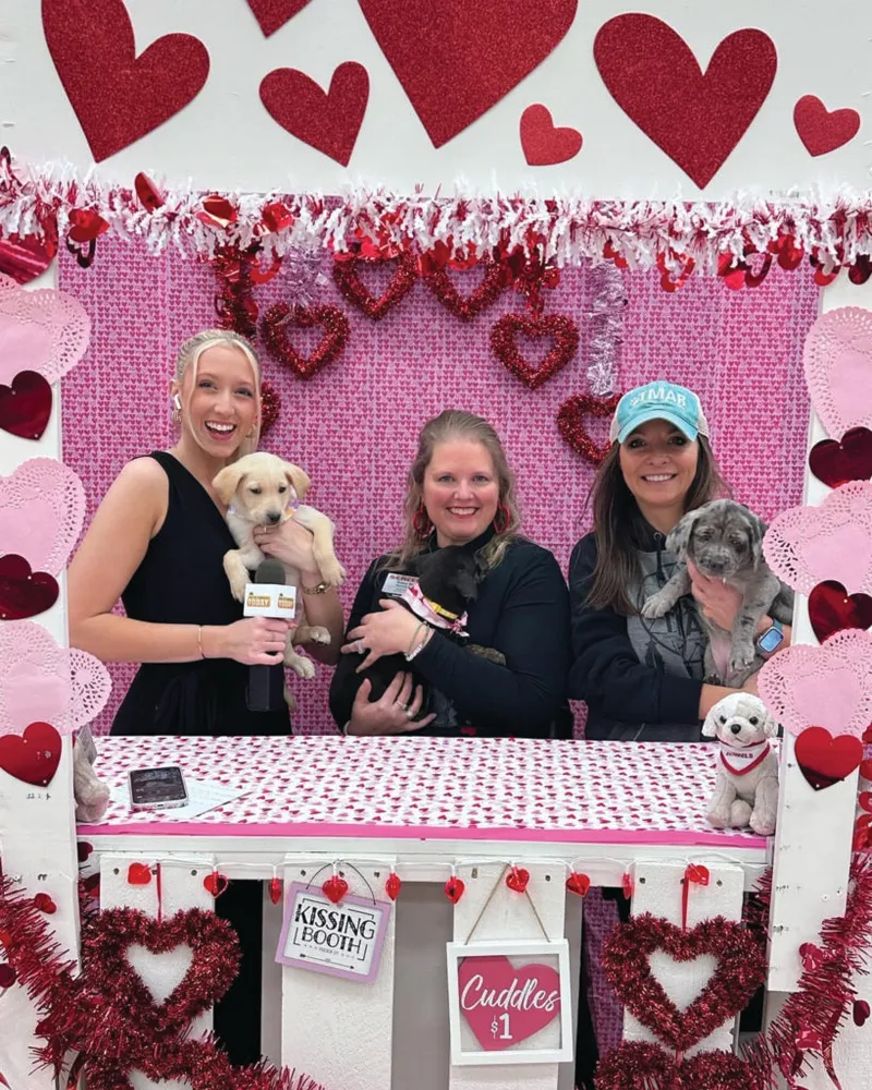 Three women holding puppies at a Valentine's Day kissing booth decorated with red hearts
