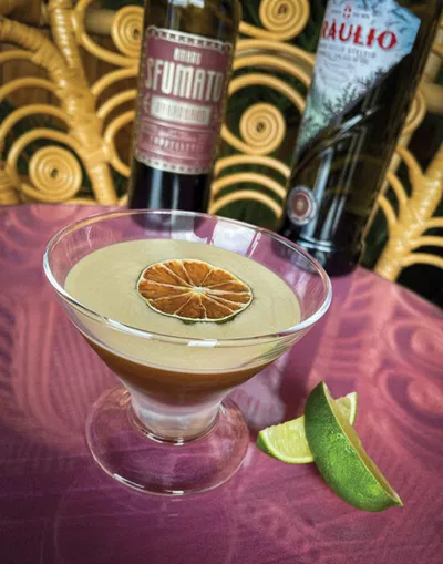 Layered cocktail in a coupe glass topped with a dried citrus wheel, with lime wedges and liquor bottles behind