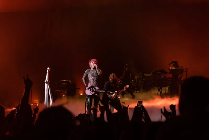 Pink-haired performer with guitar on a fog-filled stage bathed in red light, crowd cheering