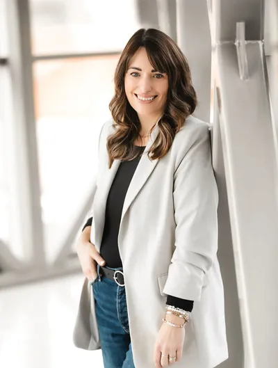 Smiling woman in a light gray blazer, black top, and jeans leaning against a white column