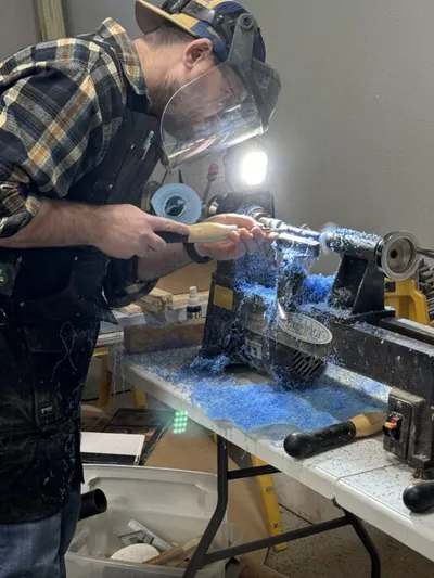 Man in face shield and flannel shirt turning a piece on a wood lathe with blue shavings flying
