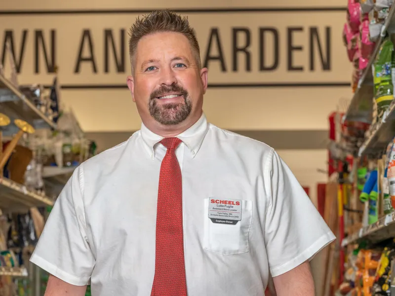 Smiling man in white shirt and red tie wearing a Scheels name badge stands in a lawn and garden store aisle