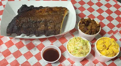 Smoked ribs on wax paper with coleslaw, baked beans, mac and cheese, and dipping sauce on a red checkered tablecloth