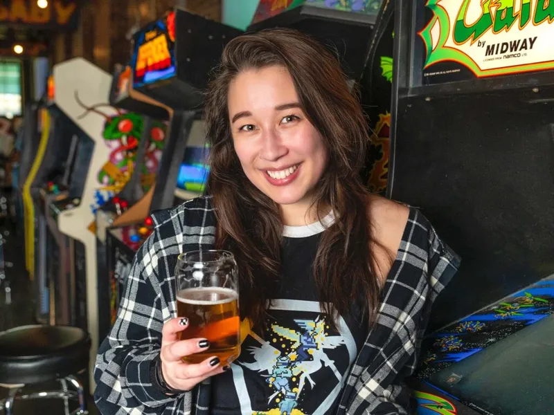 Smiling woman holding a glass of beer among vintage arcade cabinets in a bar