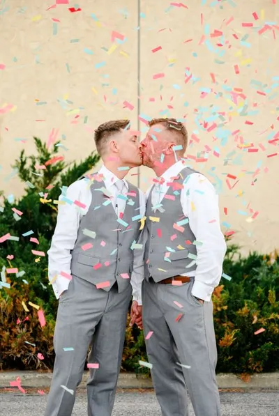 Two grooms in matching gray vests kissing amid colorful confetti