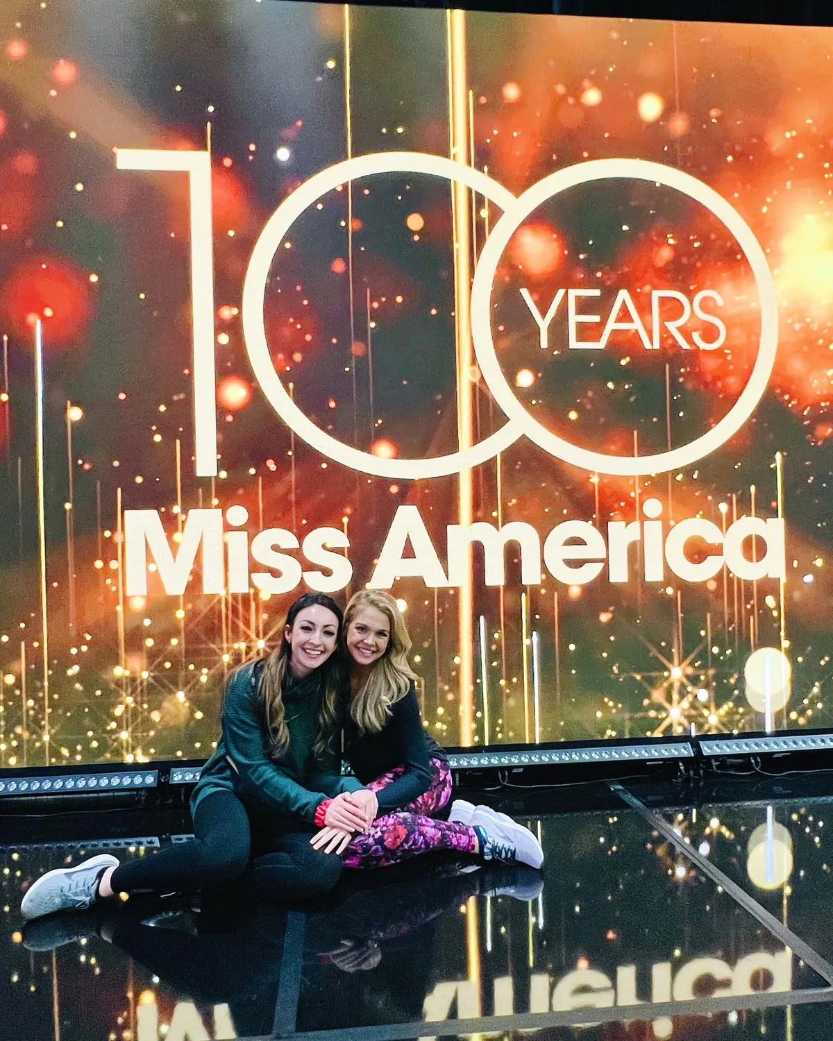 Two women sitting on a stage in front of a glowing '100 Years Miss America' backdrop