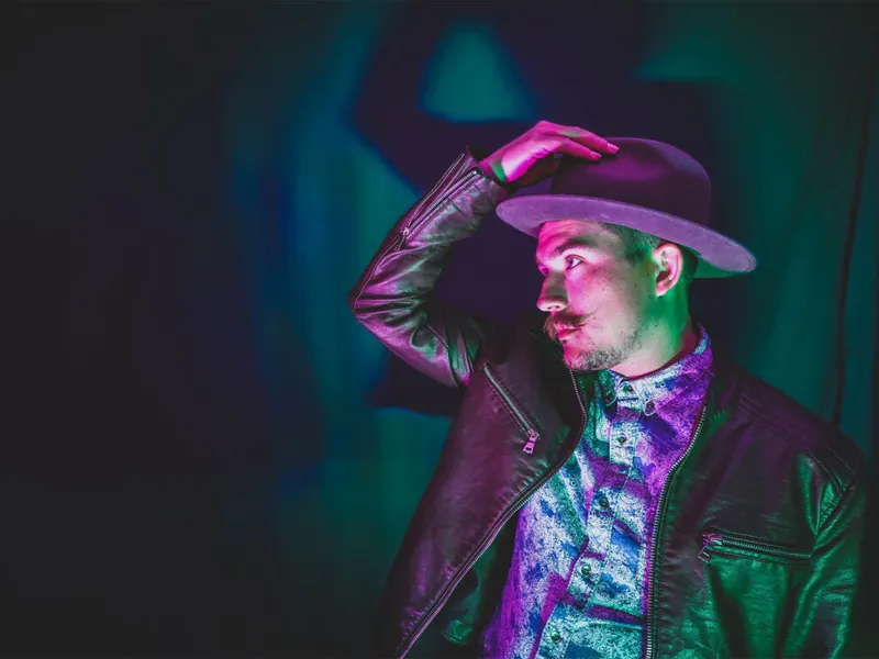 Man in wide-brim hat and leather jacket posed under purple and green stage lighting