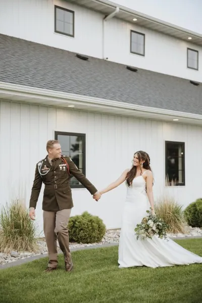 Bride in white gown and groom in military dress uniform hold hands on lawn before white barn venue