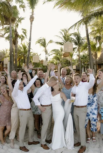 Bride and groom kiss on a sandy beach surrounded by cheering wedding guests and palm trees