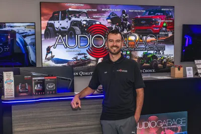 Man in black polo shirt smiling at a retail counter in front of an Audio Garage branded display