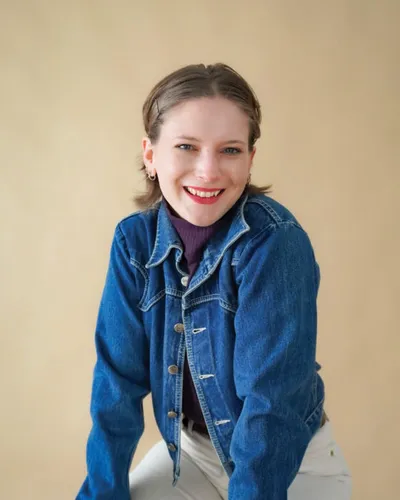 Smiling woman in a denim jacket and purple turtleneck against a beige background
