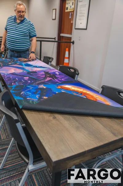 Man standing over a long table covered with a colorful fantasy-themed game mat in a meeting room