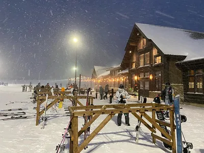 Skiers and snowboarders gather outside a lit rustic lodge during a heavy snowfall at night