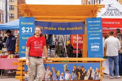 Man in red T-shirt stands smiling before a Red River Market booth at an outdoor market