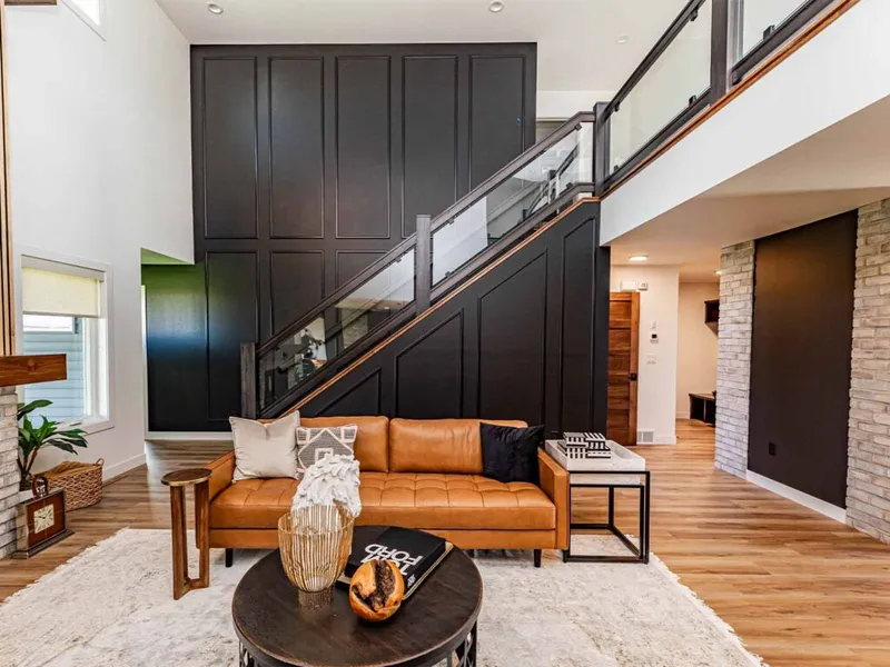 Modern living room with tan leather sofa beneath a staircase with glass railings and a dramatic black panel accent wall