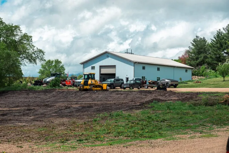 Blue metal pole barn surrounded by trucks, a yellow bulldozer, and heavy equipment on freshly graded dirt