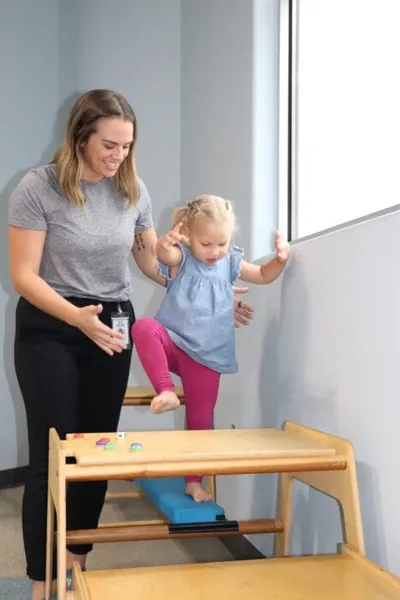 A woman steadies a toddler girl stepping onto a low wooden therapy platform