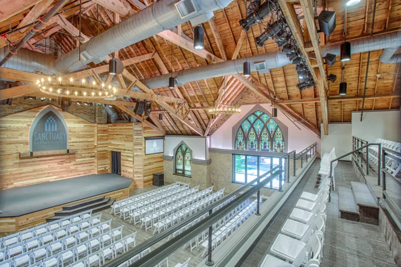 Renovated church interior with exposed wood trusses, stained glass windows, stage, and rows of white chairs
