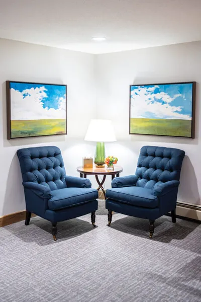 Two tufted navy armchairs flanking a round side table with lamp, beneath paired landscape paintings