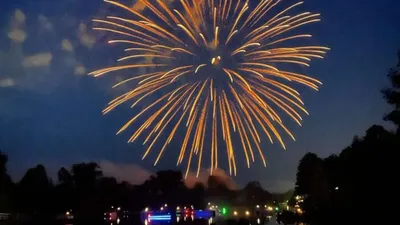 Golden fireworks burst over a lake at dusk with glowing boat lights below