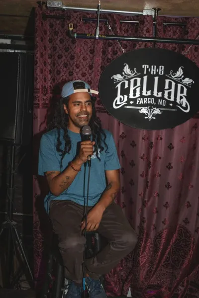 Smiling comedian with long dreadlocks and backward cap holds mic on stage at The Cellar, Fargo ND