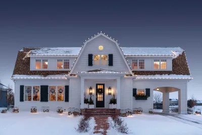White gambrel-roof house decorated with string lights and wreaths in winter snow at dusk