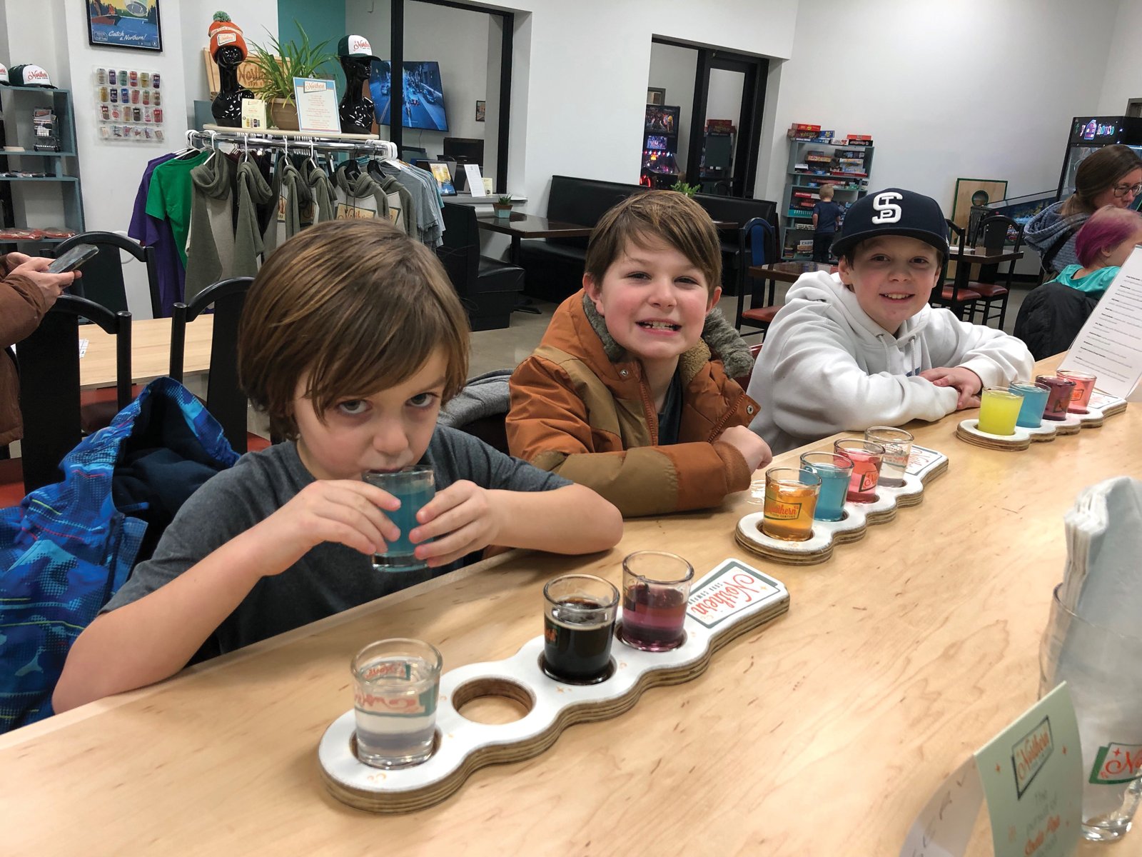 Three kids smile at a counter with flights of colorful soda samples