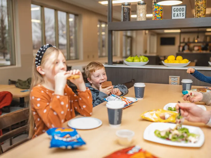 Two children eating sandwiches and snacks at a cafeteria table with a 'Good Eats' sign in the background