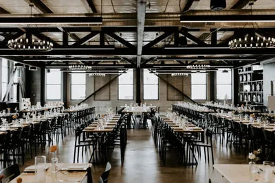 Industrial event hall with long banquet tables, black chairs, and chandeliers under exposed steel trusses