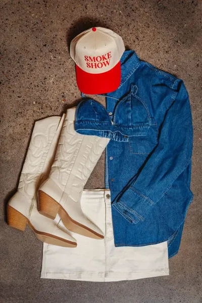 Flat lay of white cowboy boots, denim shirt, mini skirt, and a red 'Smoke Show' trucker hat