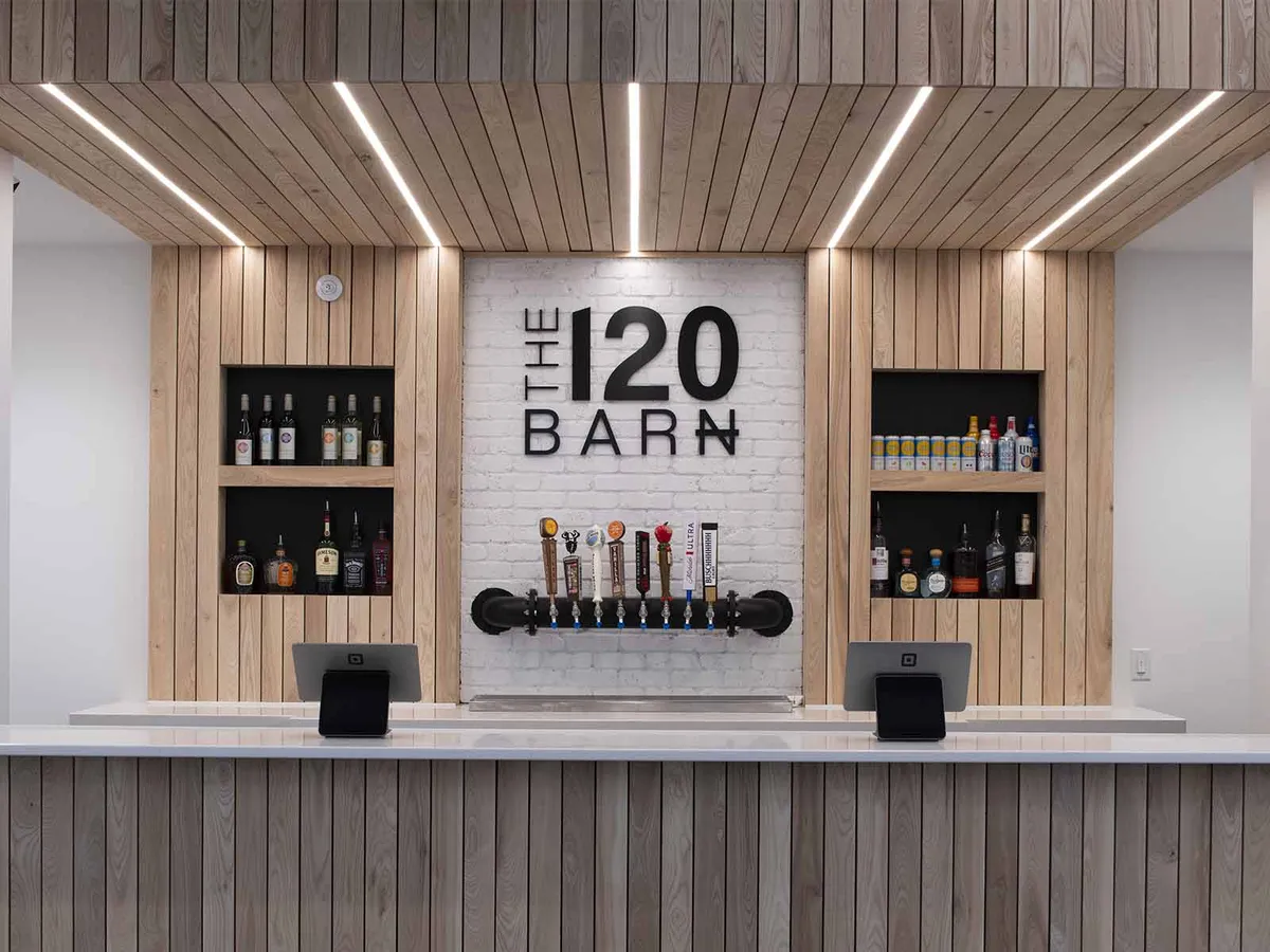 Bar interior with wood-slat walls, draft taps, liquor shelves, and a 'The 120 Barn' sign on white brick