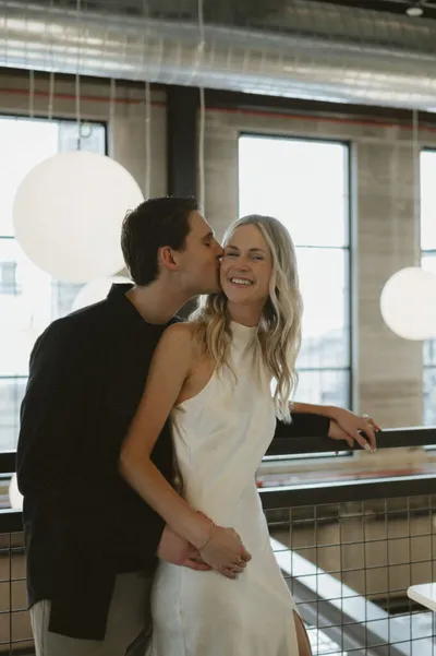 Smiling woman in white dress being kissed on the cheek by a man in a dark jacket inside a modern restaurant