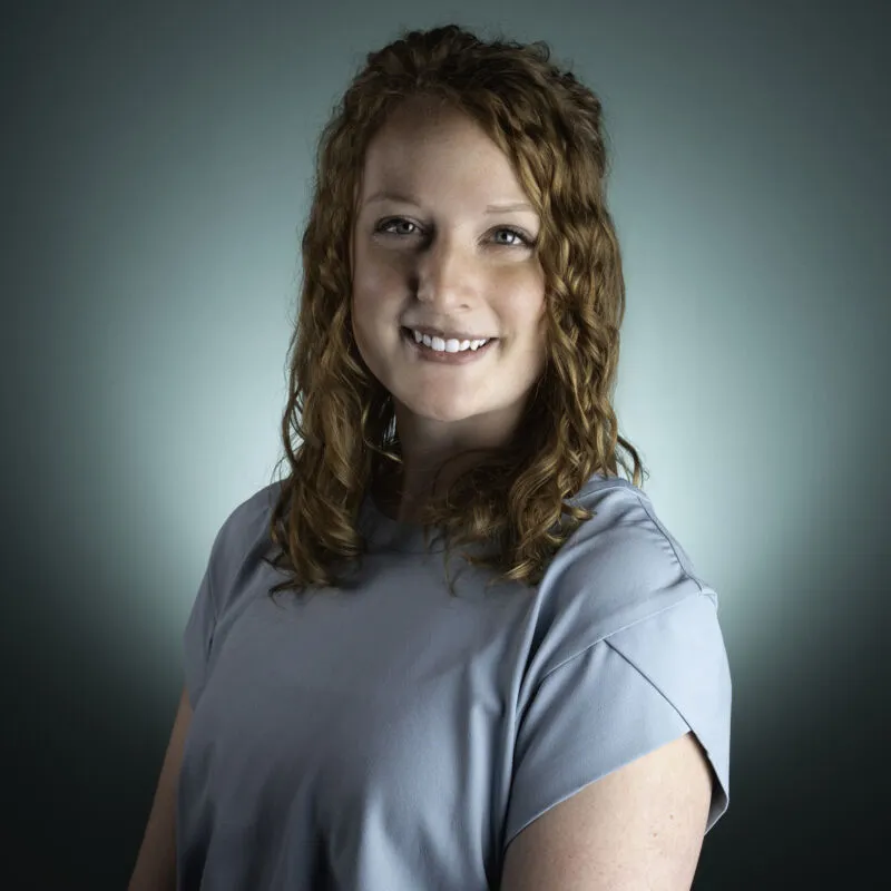 Smiling woman with curly auburn hair wearing a light blue top against a teal backdrop