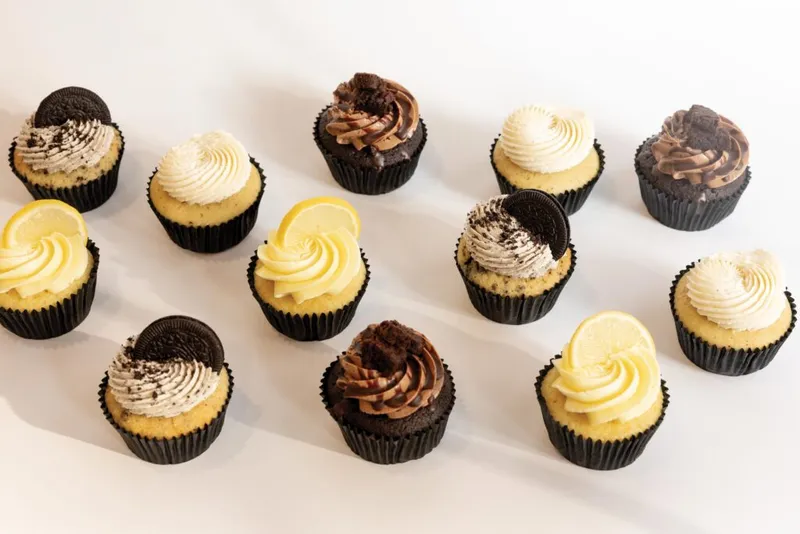 Assorted cupcakes with swirled frosting, lemon slices, chocolate toppers, and cookie garnishes on a white surface
