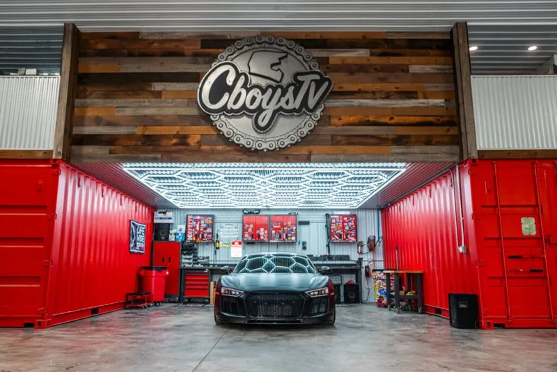 Dark sports car parked inside a red shipping-container garage beneath a CboysTV logo sign