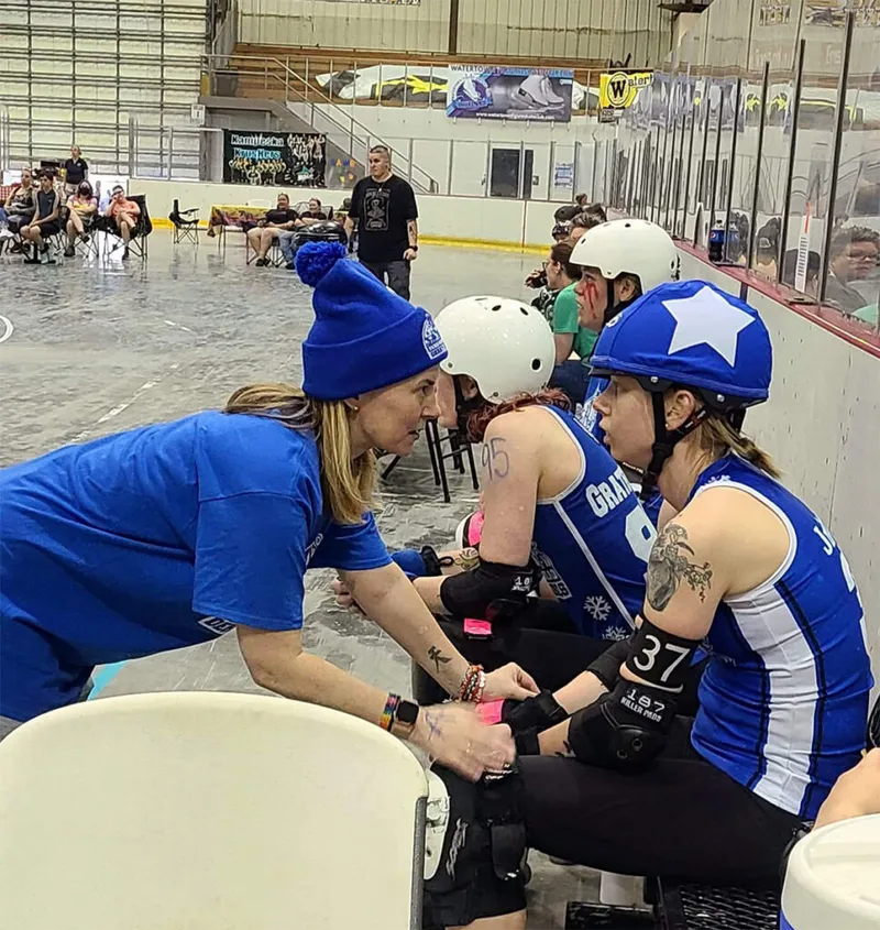 A coach in a blue beanie leans in to talk with two roller derby skaters in helmets on the bench