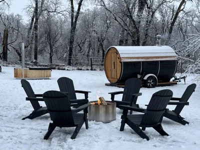 Black Adirondack chairs circling a fire pit in snow, with a barrel sauna on a trailer behind