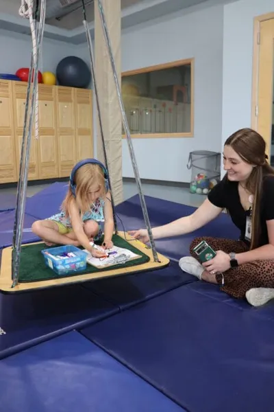 Young girl sitting on a platform swing doing an activity while a smiling therapist looks on in a sensory gym