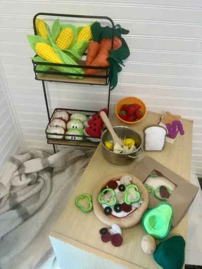 Felt play food including corn, carrots, pizza, and vegetables arranged on a small wooden table and tiered rack