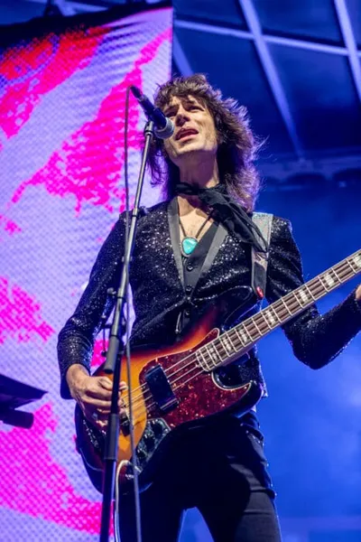 Long-haired guitarist in a black sequin jacket performs at a mic on a lit stage