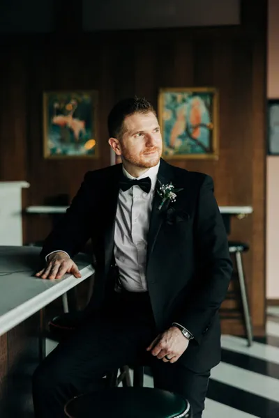 Man in black tuxedo with boutonnière seated at a bar, gazing to the side