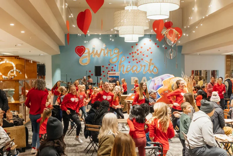 Crowd in red Giving Hearts Day shirts dancing beneath paper heart decorations in a festive indoor venue