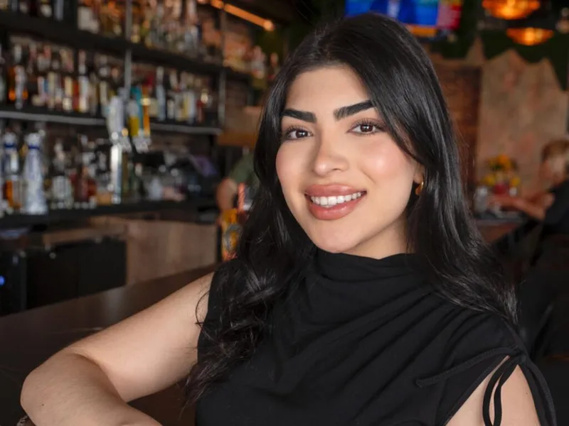 Smiling woman with long dark hair in a black top seated at a bar with shelves of bottles behind her