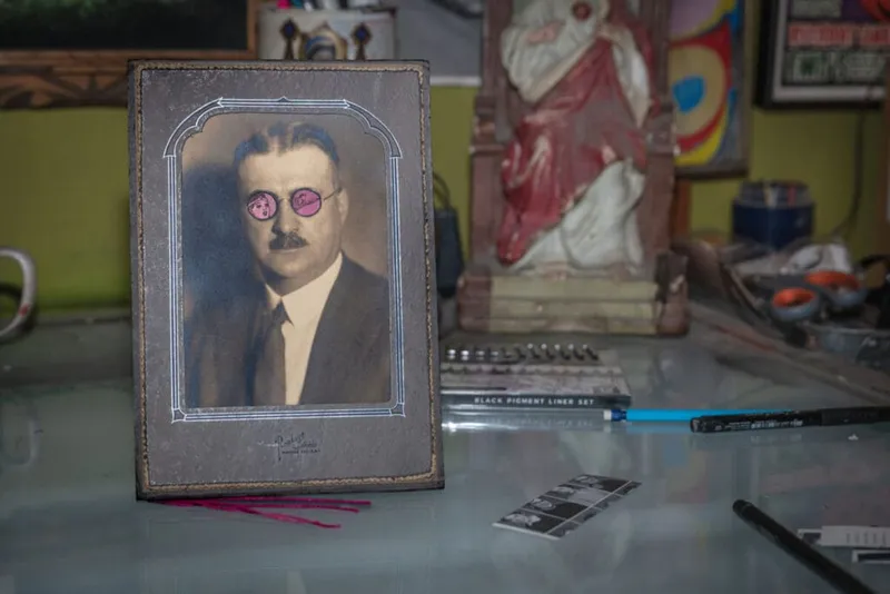 Vintage framed portrait altered with drawn pink glasses, on a cluttered artist's desk with pens and sketchbook