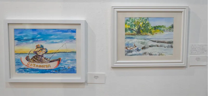 Two framed watercolor paintings on a white gallery wall, one depicting a cat fishing from a boat labeled "CATamaran