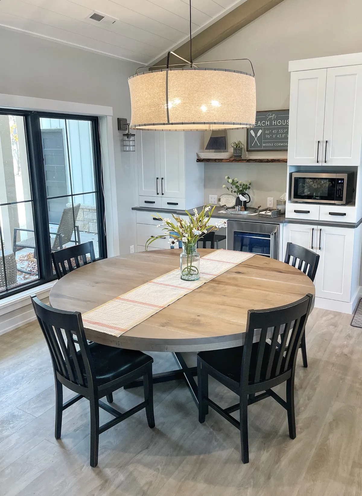 Round wood dining table with black chairs and floral centerpiece beneath a drum pendant light